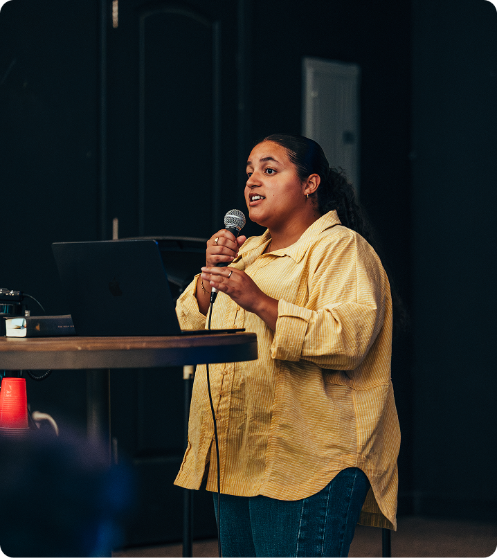 Person speaking into a microphone beside a laptop at a podium in a dark conference room