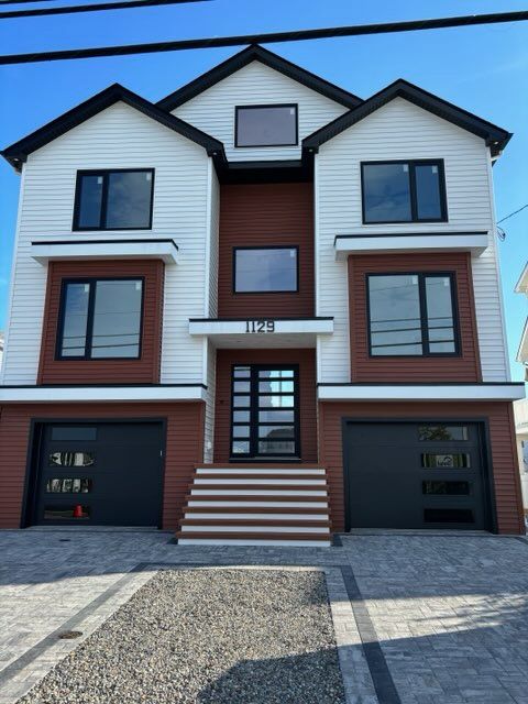 Modern three-story house with white siding, brown trim, and two garage doors under a clear blue sky