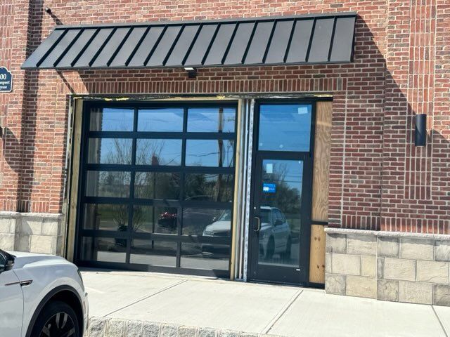 Brick storefront entrance with black awning, glass doors, and a parked white car at left