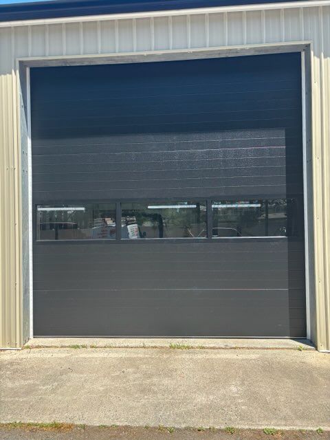 Closed dark gray industrial roll-up door on a beige building, with a narrow window strip across the middle.
