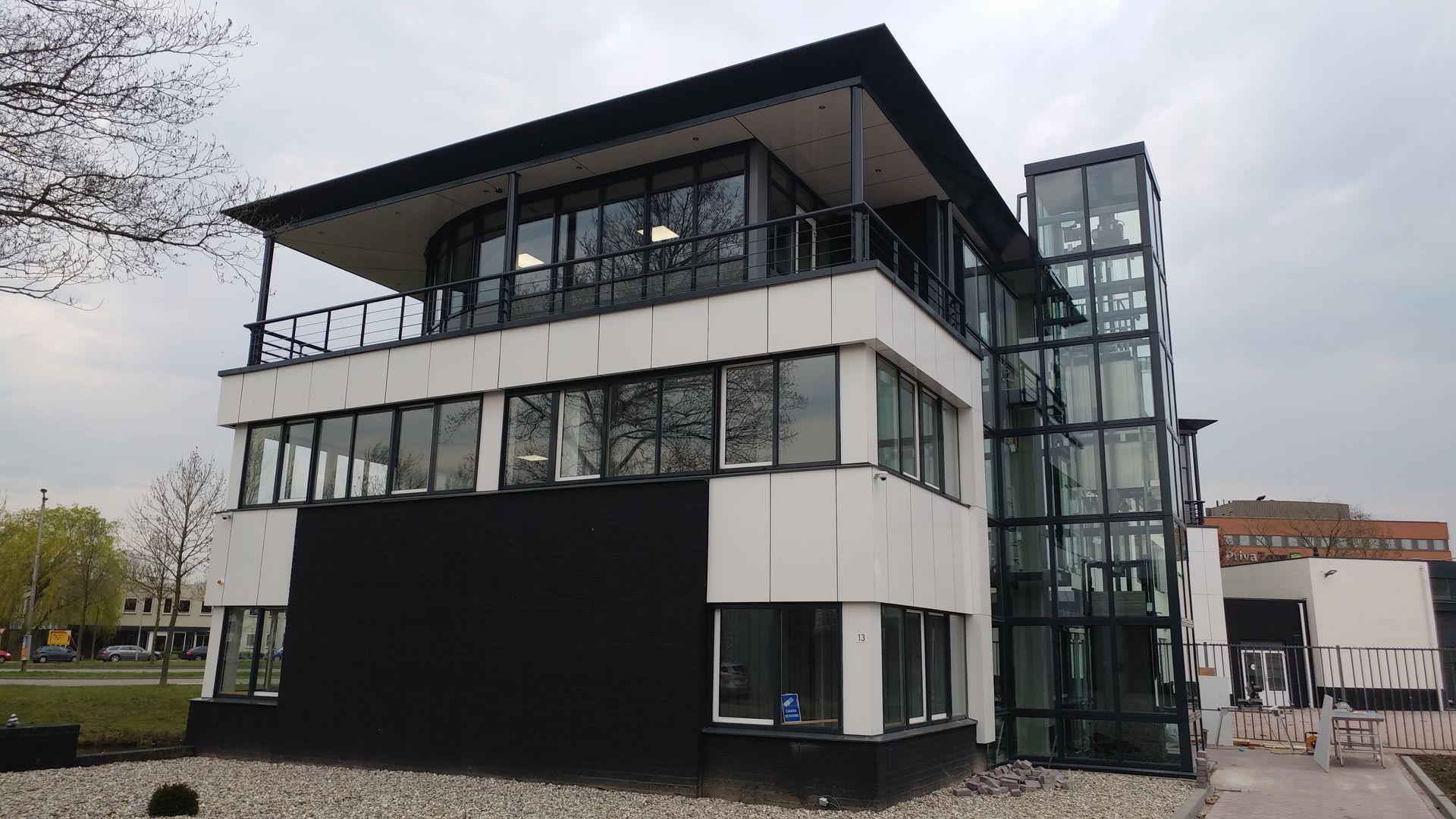 Modern white and black building with glass elevator and outdoor balcony under overcast sky.
