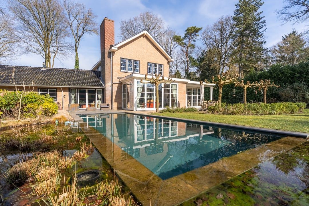 House with rectangular pool, lush green lawn, tall trees, and a brick chimney.