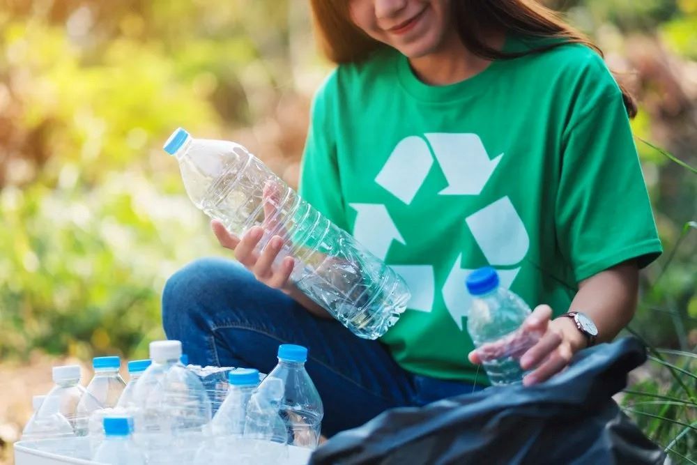 A Woman in a Green Shirt is Sitting on the Ground Holding a Plastic Bottle — The Territory Can Man In Winnelie, NT