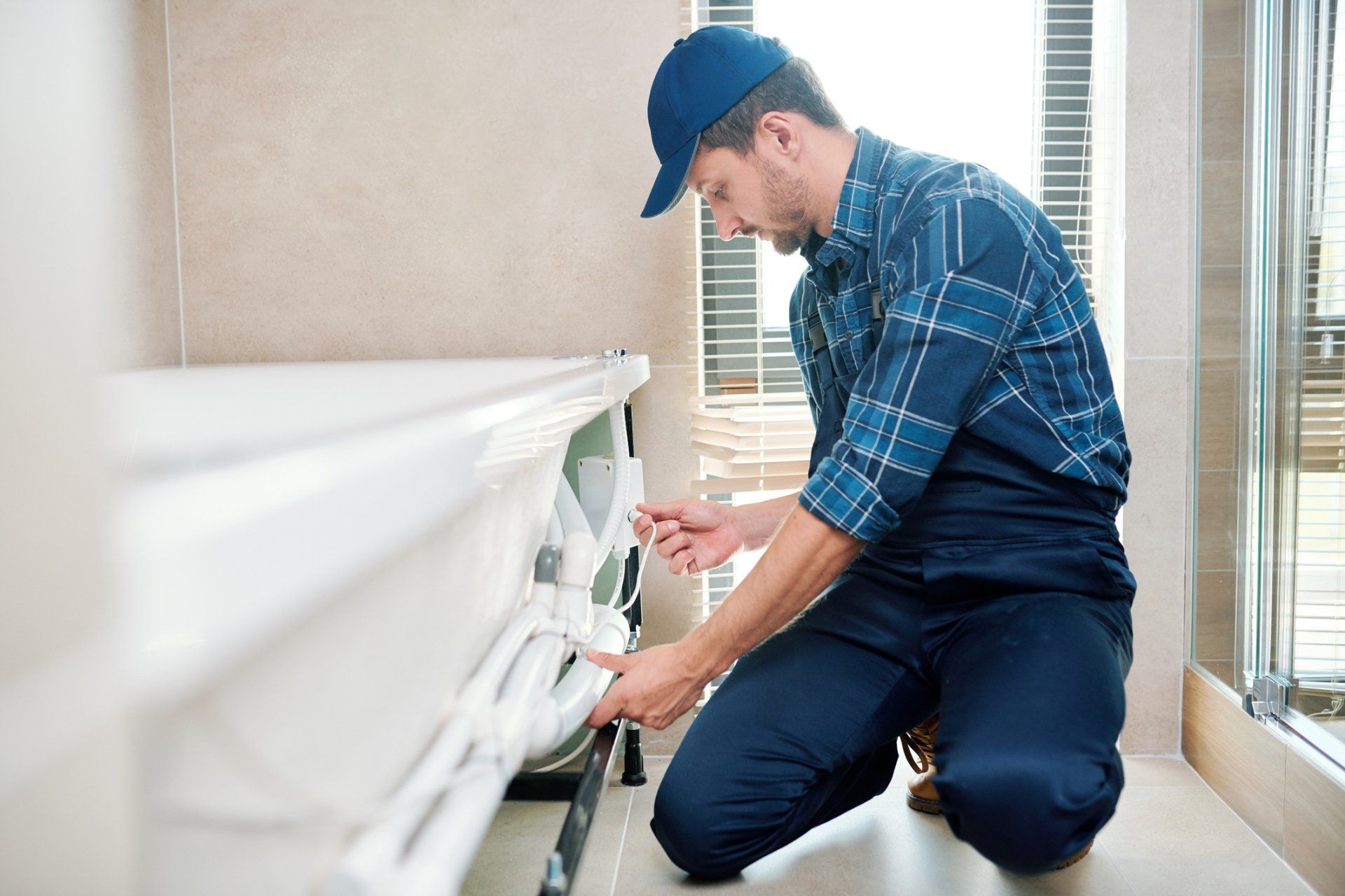 Plumber in blue cap and plaid shirt repairs bathtub in bathroom.