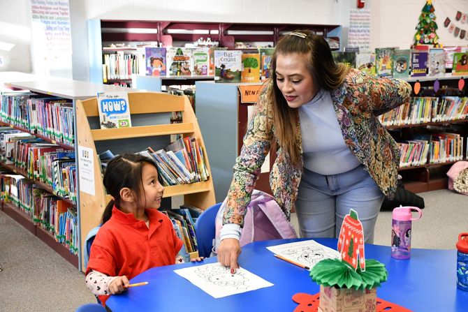 Woman and child at table in a library. Woman points at paper. Child looks up. Bookshelves in background.