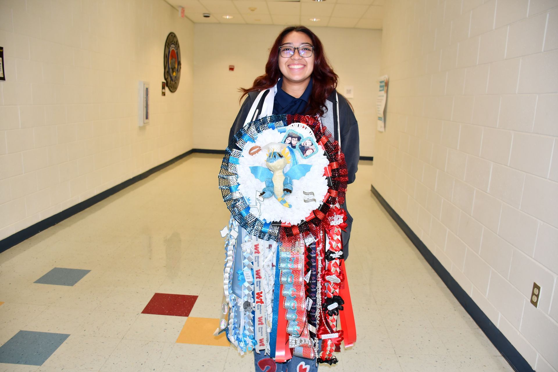 Female student wearing blue and red homecoming mum smiles in school hallway.