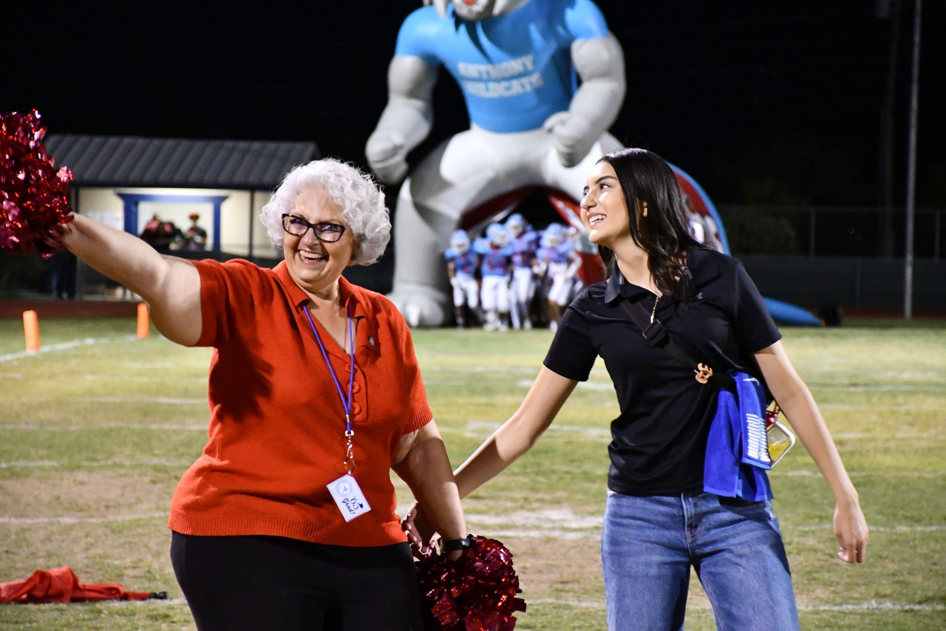 Woman wearing red shirt and holding pom-pom waves at crowd while student reaches and smiles towards her.
