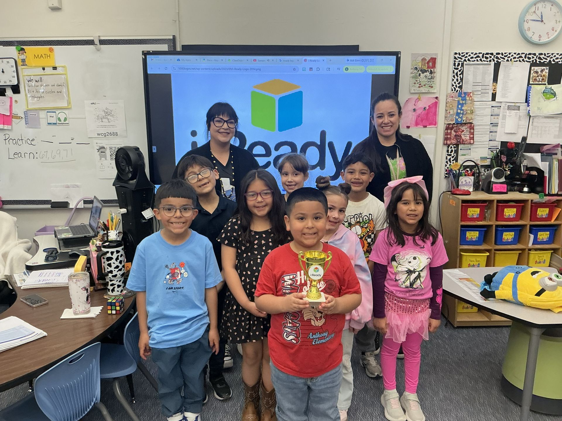 Five children and two adults pose in a classroom in front of a large screen and colorful cubbies.
