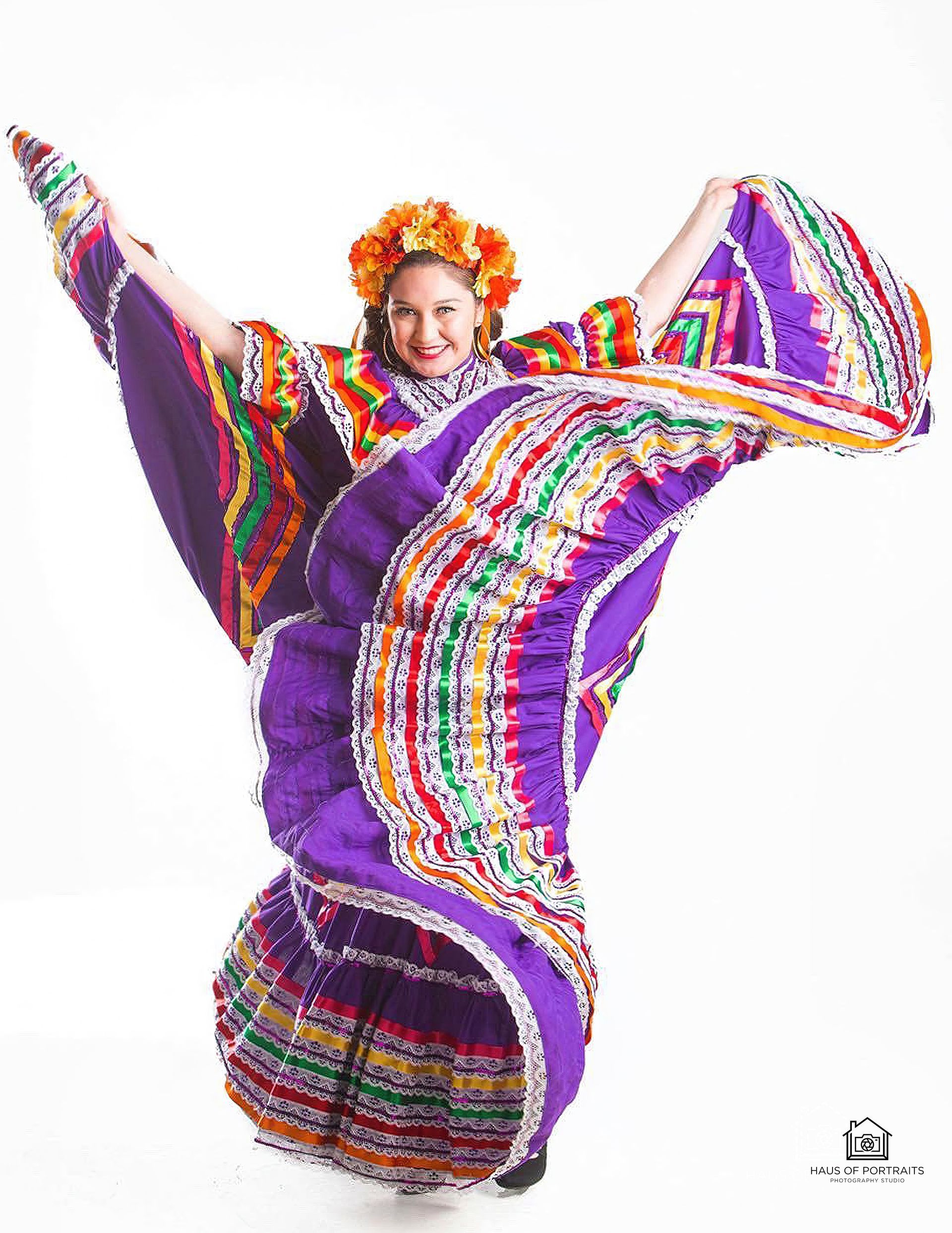 Person in a vibrant purple, multicolored folkloric dress, arms raised in a dynamic dance pose.