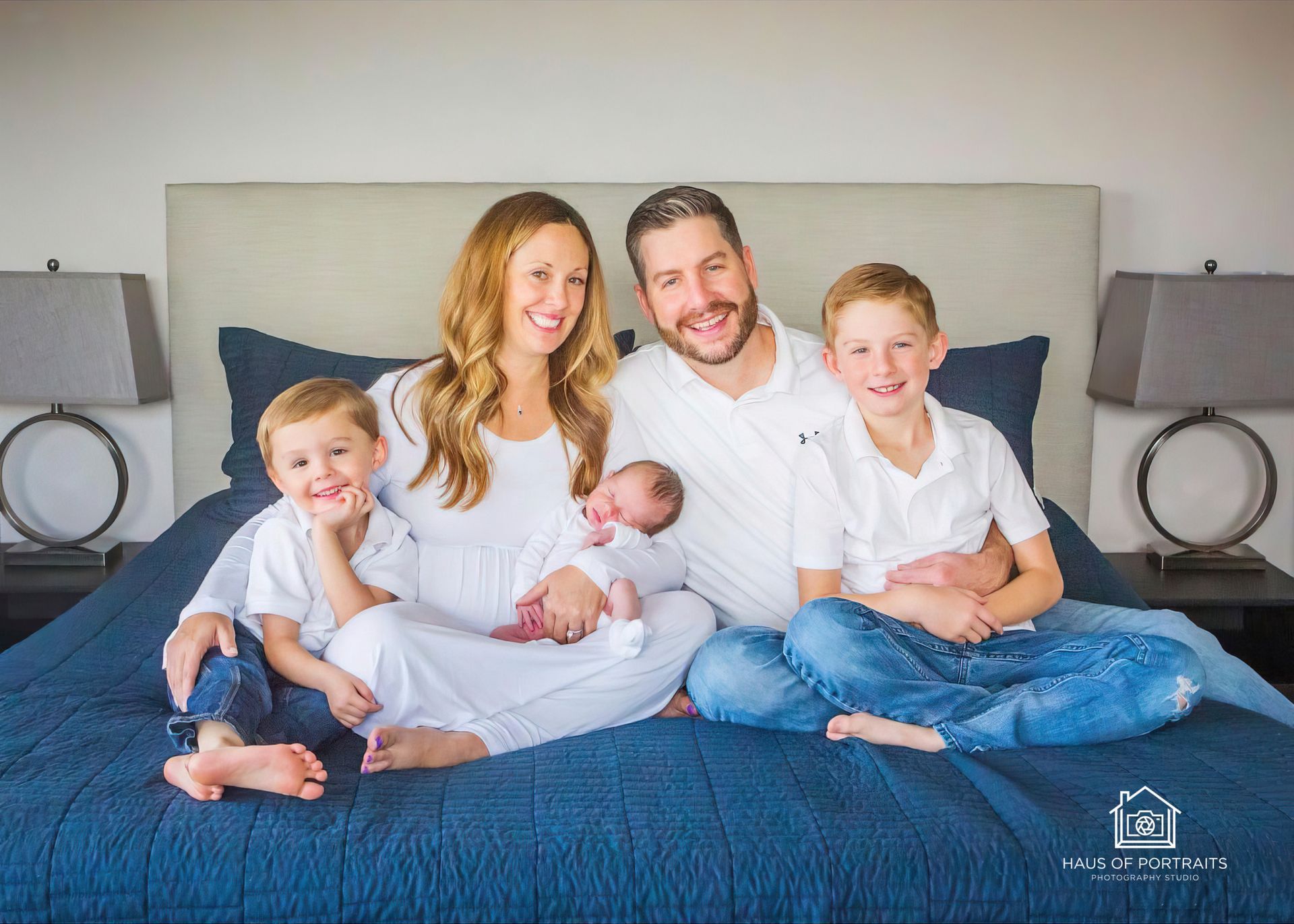 Family of four with a newborn seated on a bed in a bright bedroom, smiling at the camera.