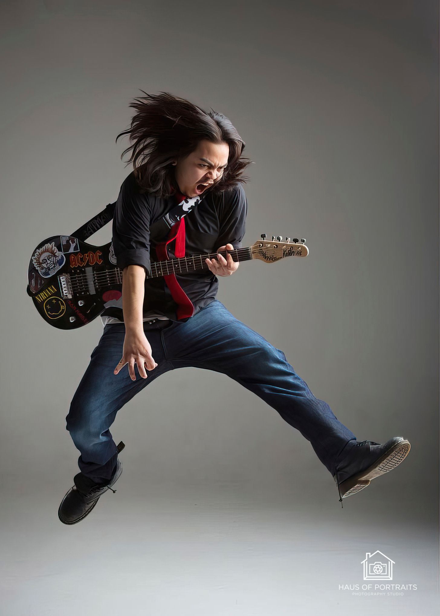 Person mid-jump with electric guitar, long hair flying, wearing a black jacket, red tie, and blue jeans.
