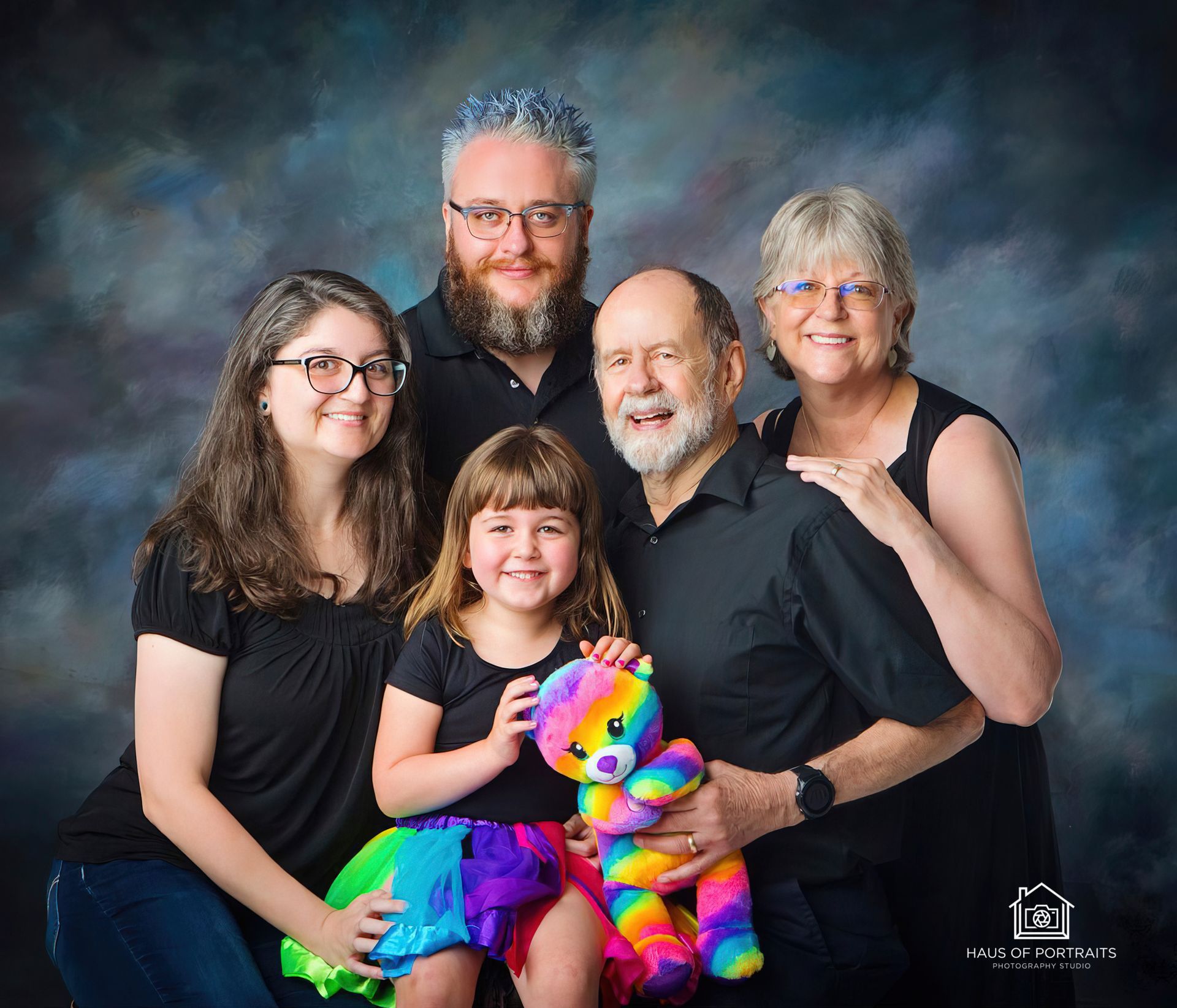 Five smiling people pose in front of a blue backdrop, with a colorful stuffed toy in front.