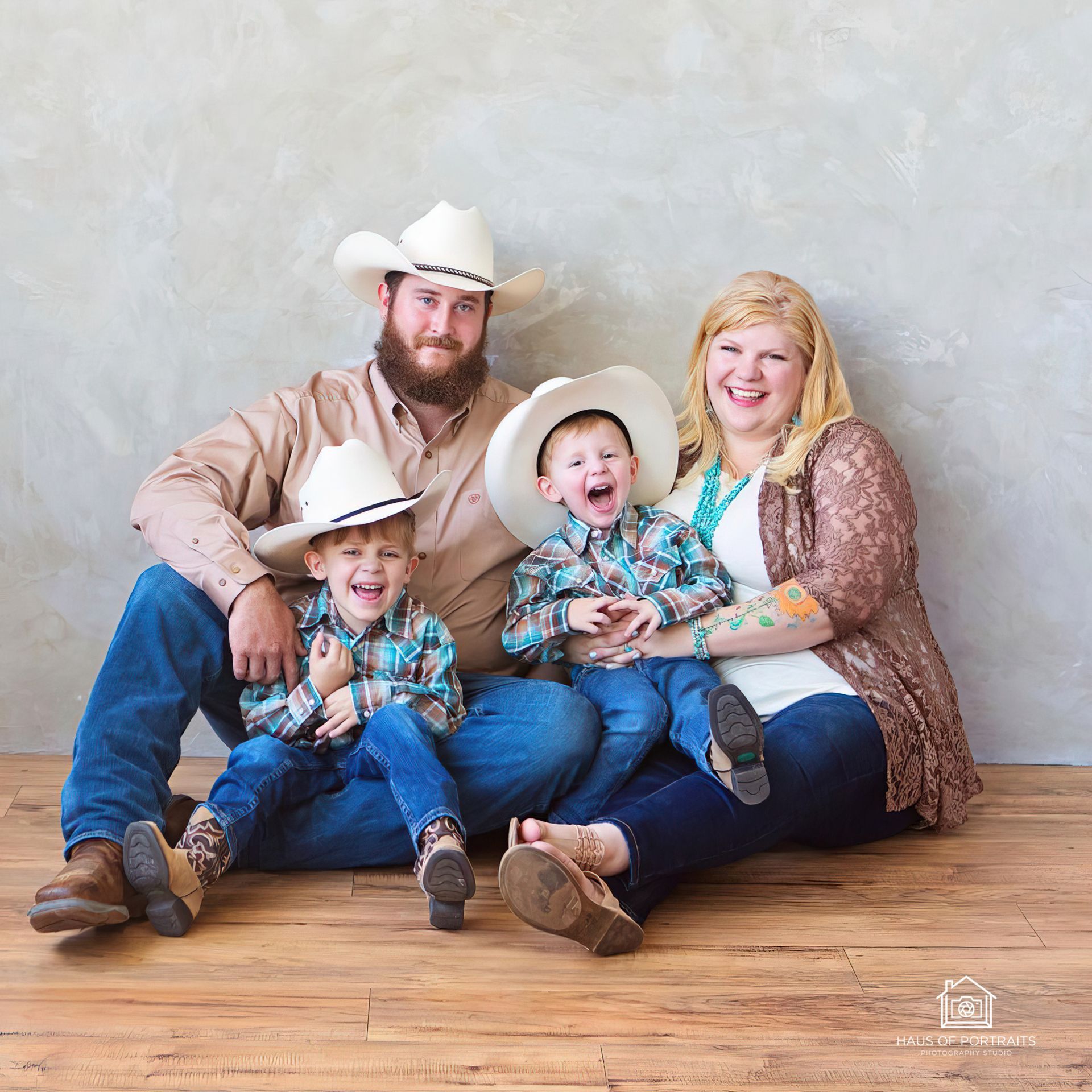 Family of four seated on a wooden floor, smiling in cowboy hats against a gray studio backdrop.