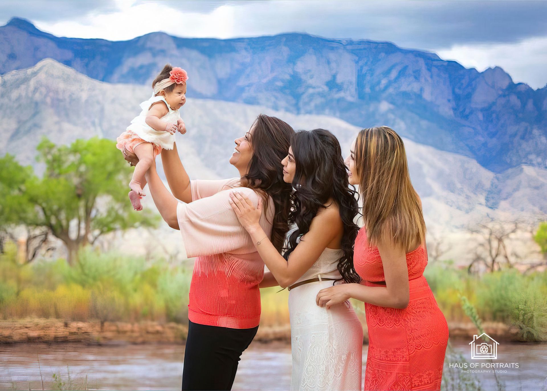 Three women holding a baby outdoors with mountains and desert plants in the background