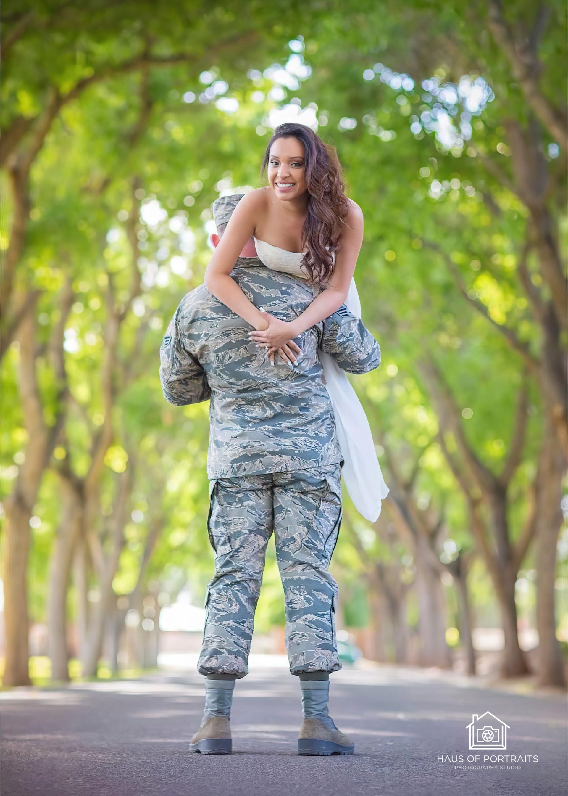 Person in military uniform giving a piggyback ride on a tree-lined path.