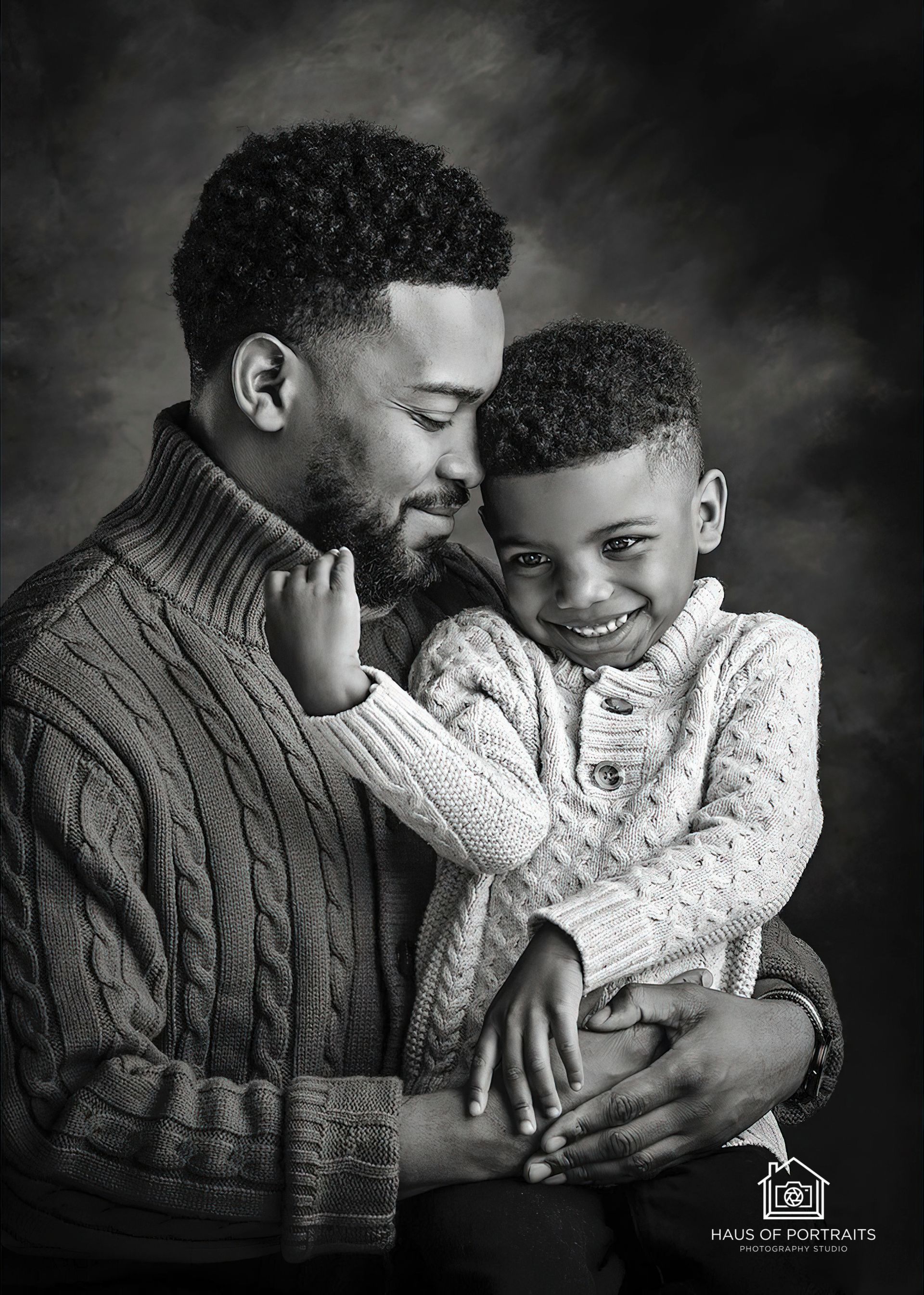 Father and child embracing in a black-and-white studio portrait, both smiling softly.