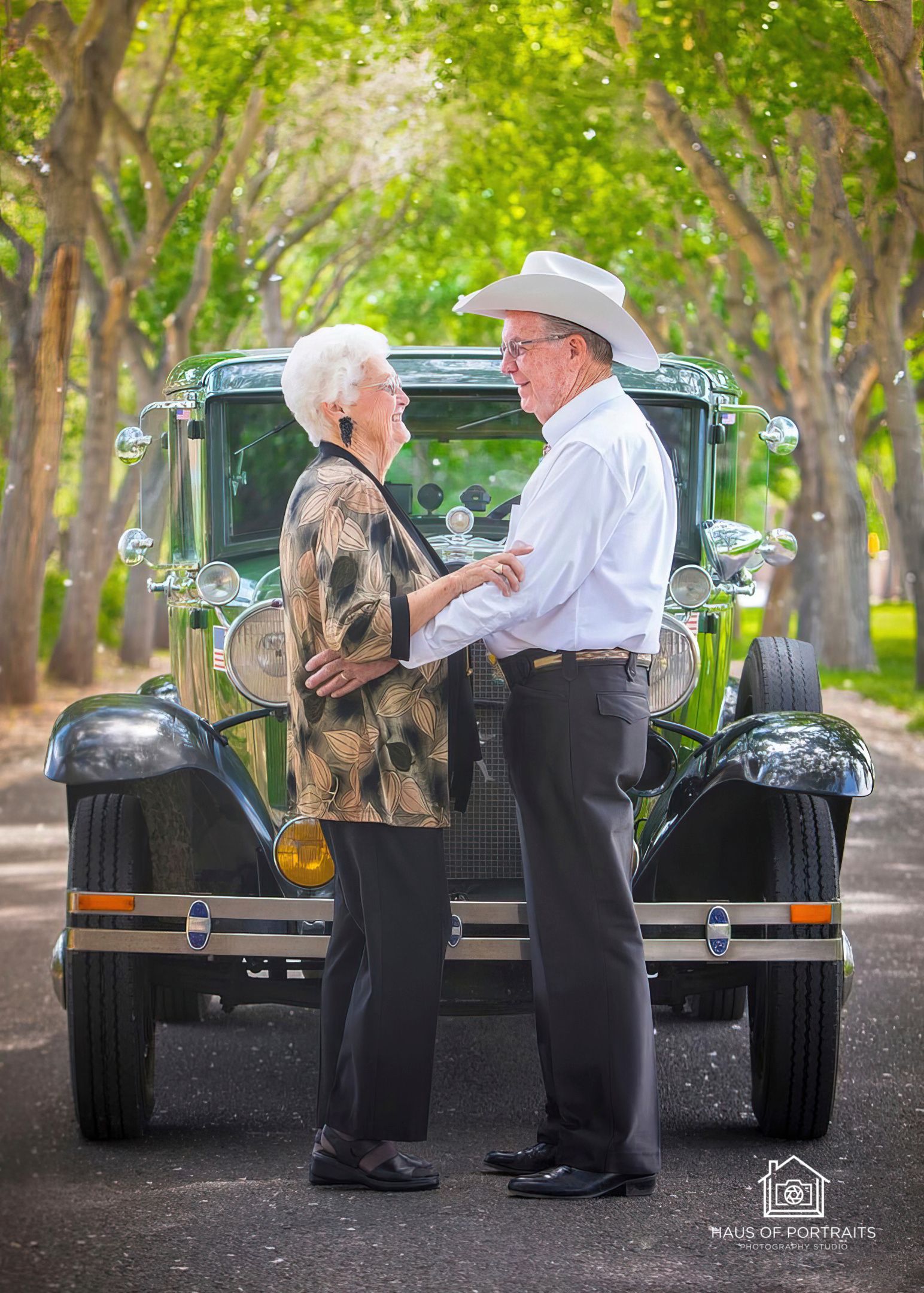 Two people standing face to face in front of a vintage car on a tree-lined road