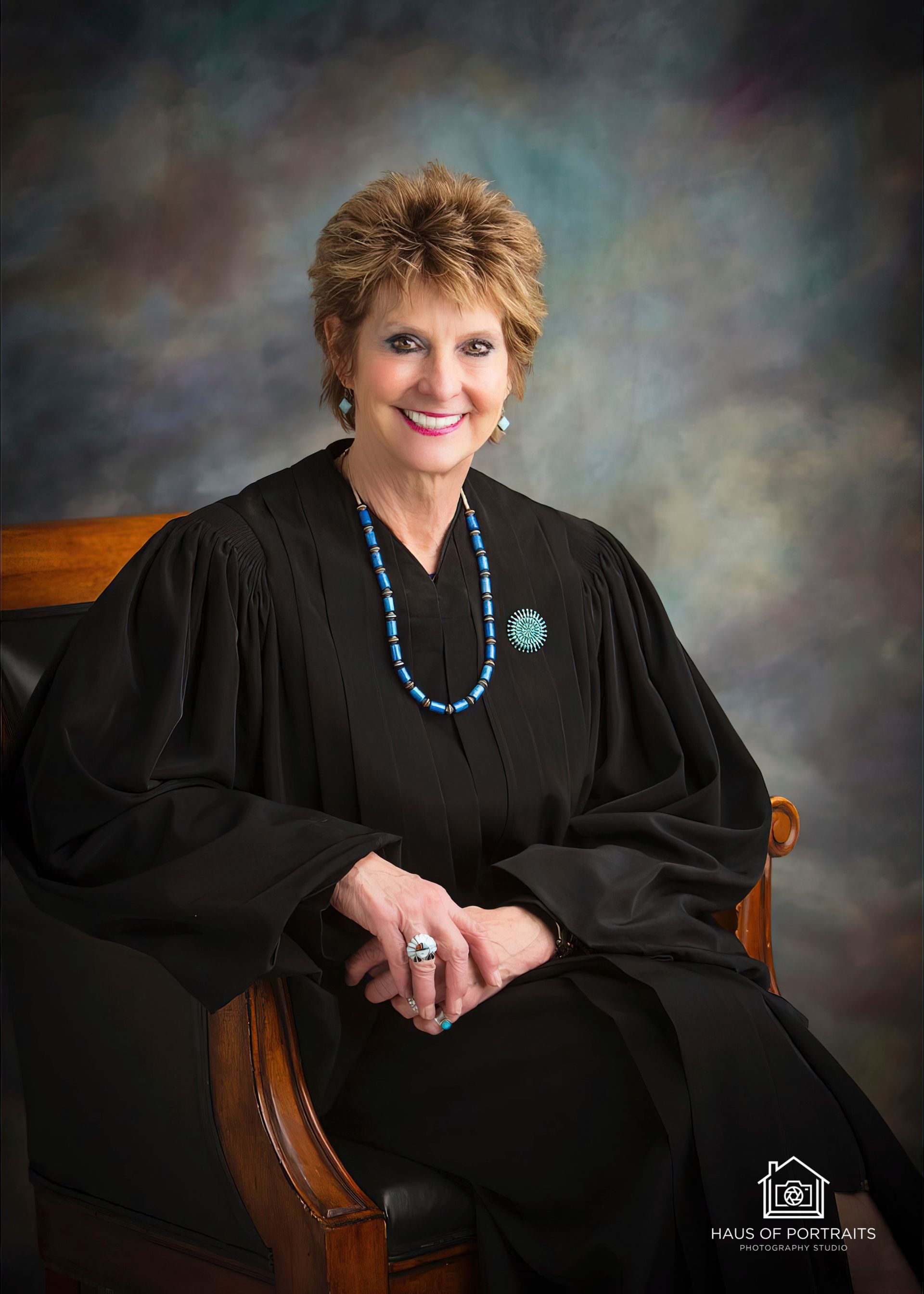 Professional portrait of a woman in a black robe, seated and smiling, with a turquoise necklace against a studio backdrop.