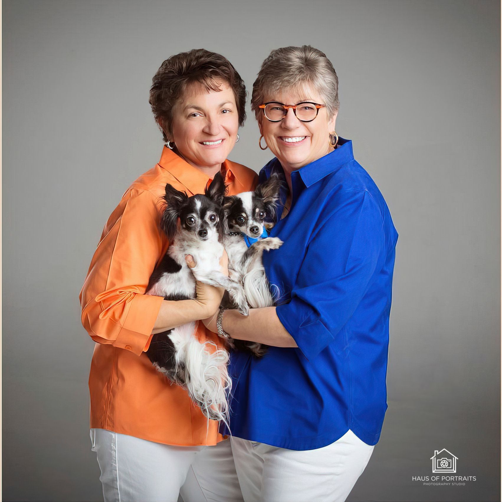 Two smiling adults holding a small black-and-white dog against a gray studio backdrop