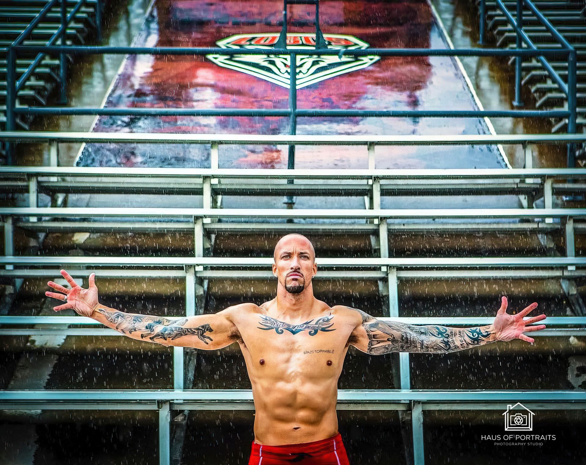 Tattooed shirtless man with arms outstretched on stadium bleachers beneath a colorful mural