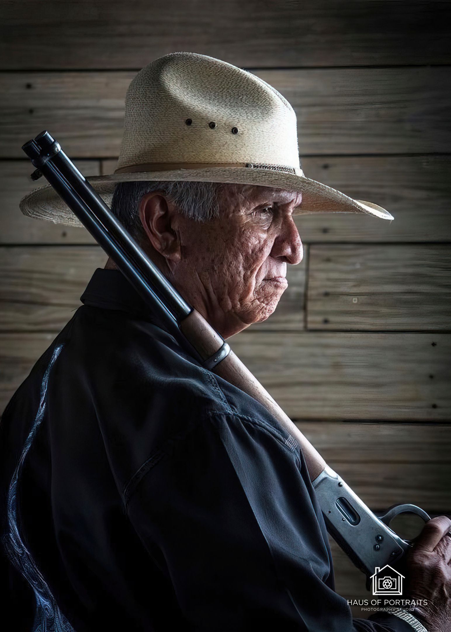Profile of a man in a straw hat holding a rifle, standing against a wooden wall