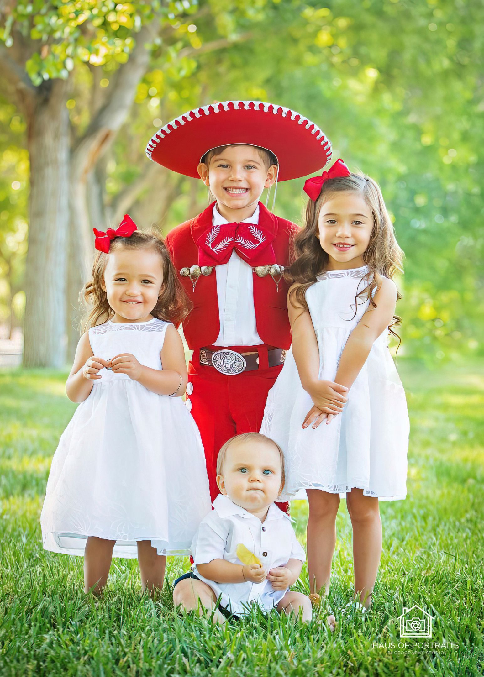 Four children in white and red outfits posing on grass outdoors, with one wearing a red sombrero.