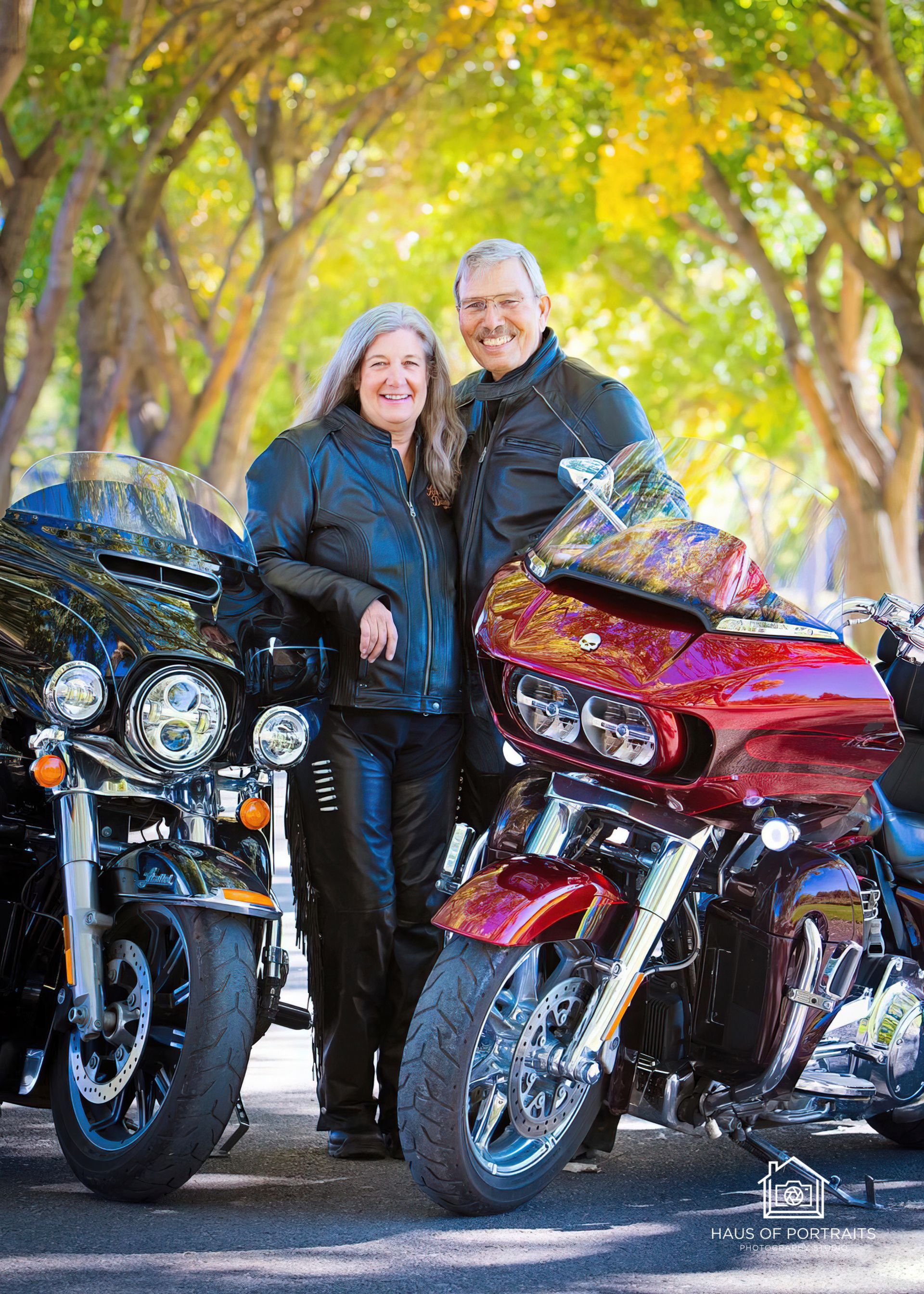Two people standing between a black and a red motorcycle on a tree-lined road.