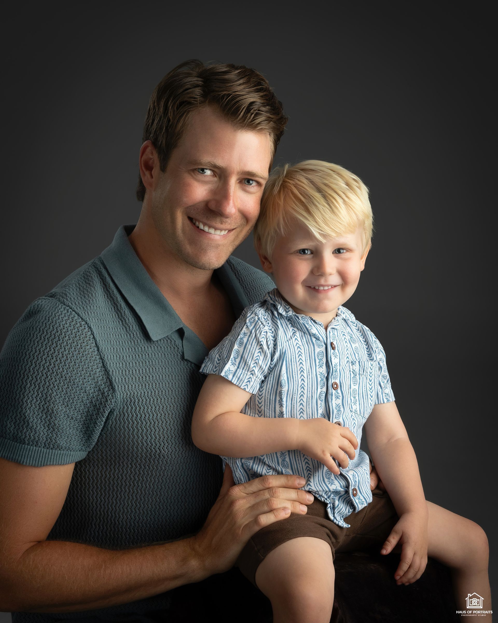 Smiling man and young child seated together against a dark studio backdrop