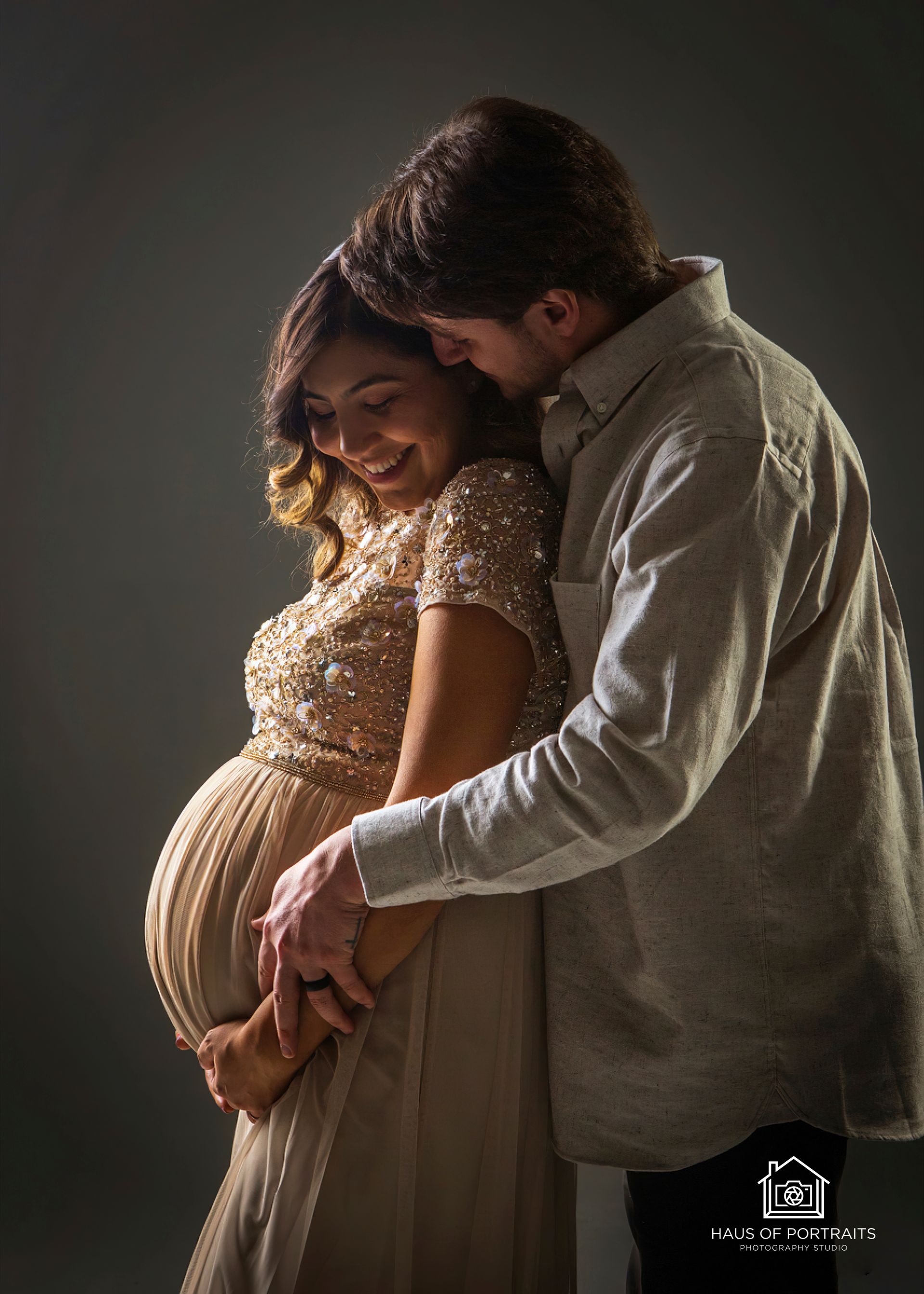Pregnant woman smiling as partner embraces her from behind against a dark studio backdrop