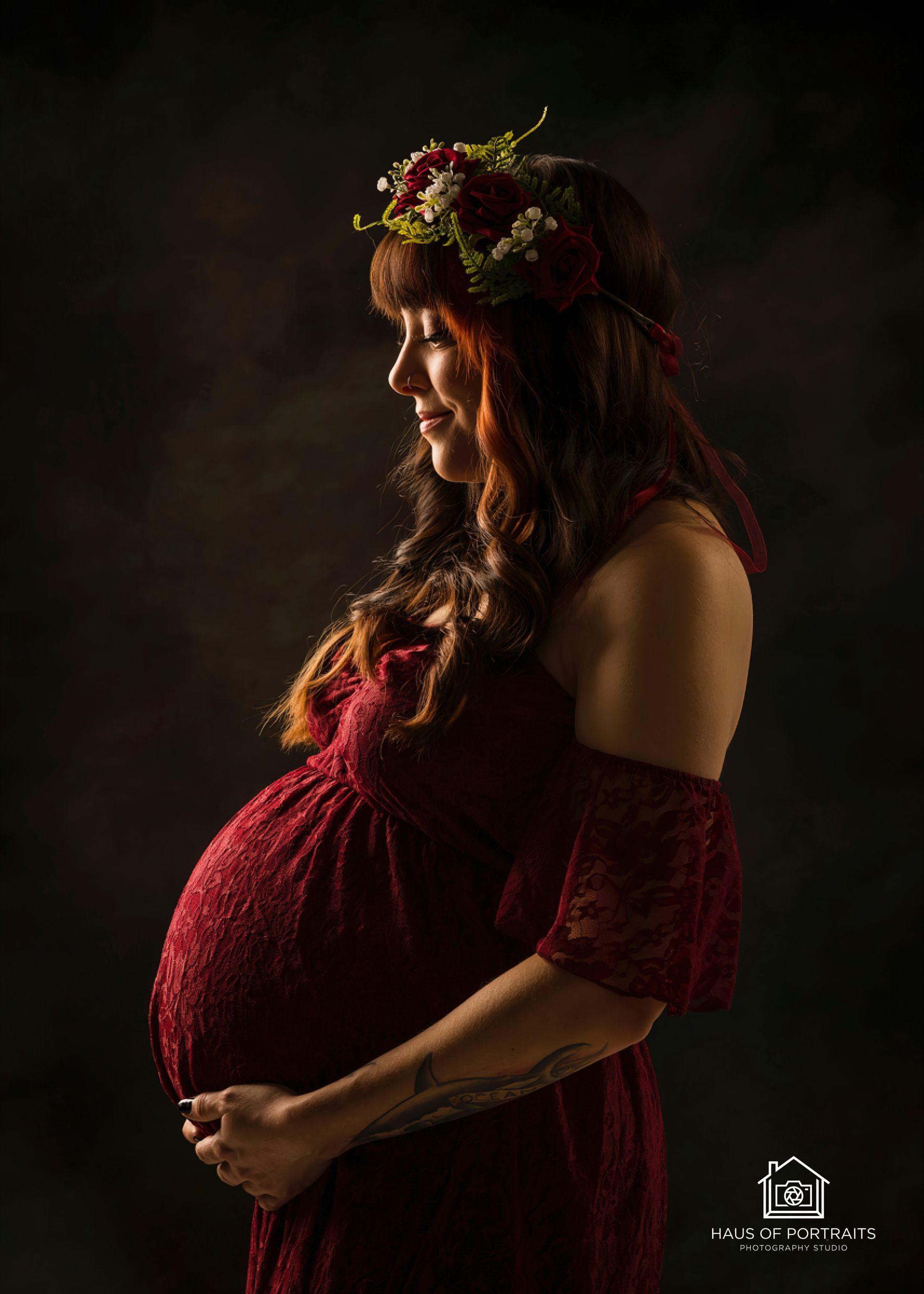 Pregnant woman in a red off-shoulder dress, standing in profile with a floral crown against a dark background