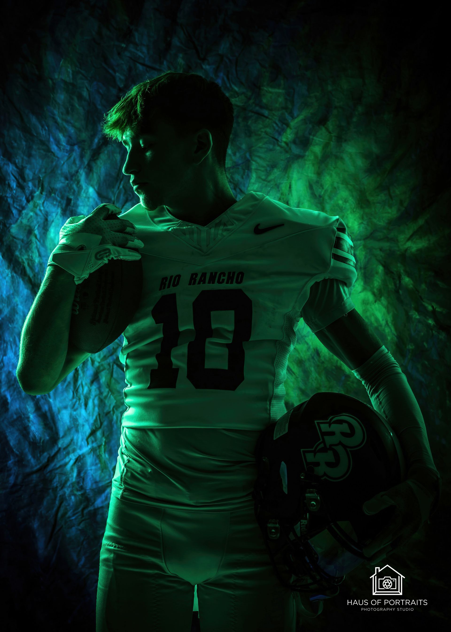 Green-lit football player in uniform holding a helmet, posed against a smoky dark background.