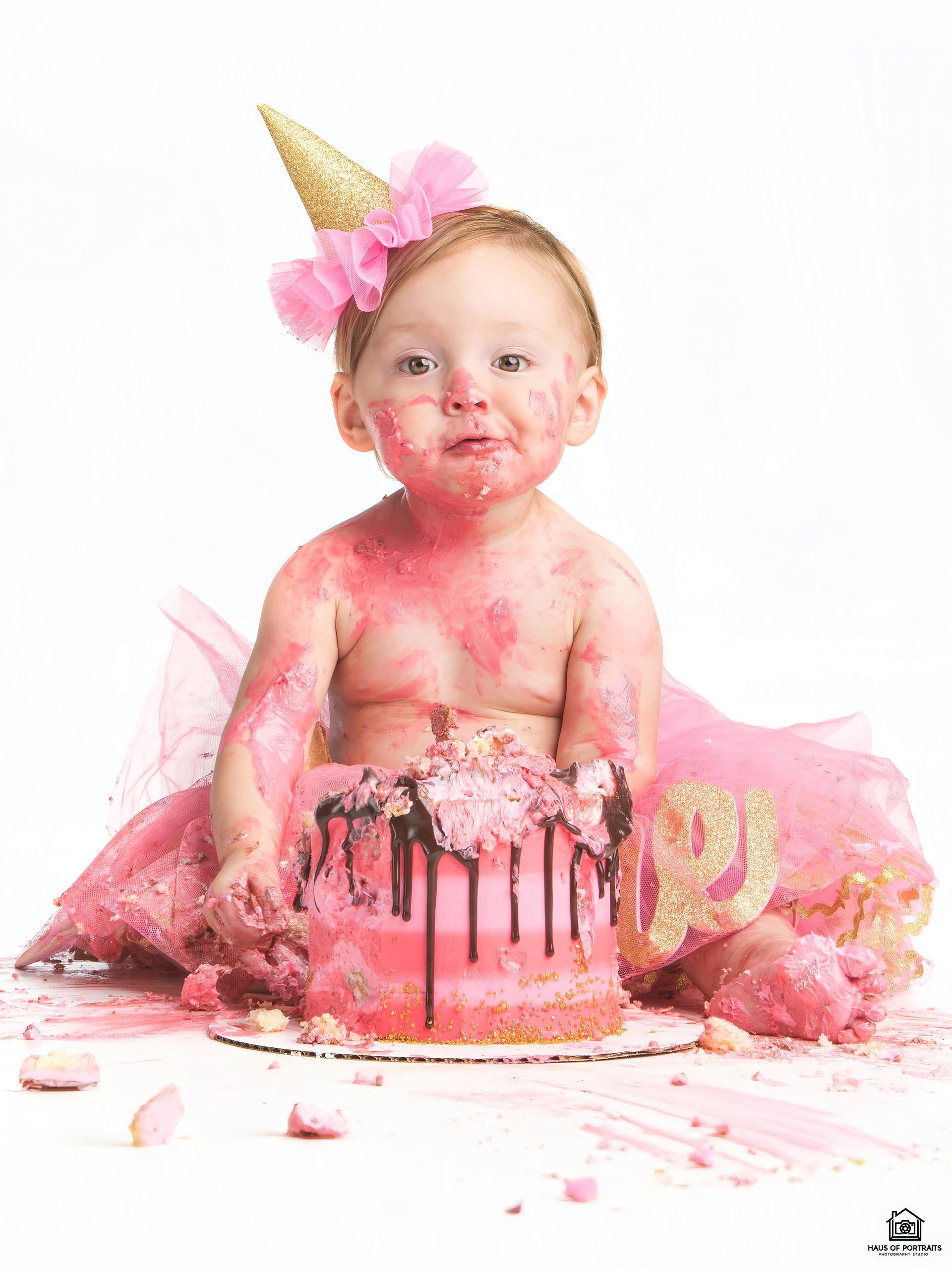 Baby sitting beside a pink cake, covered in frosting with a gold party hat and pink tutu