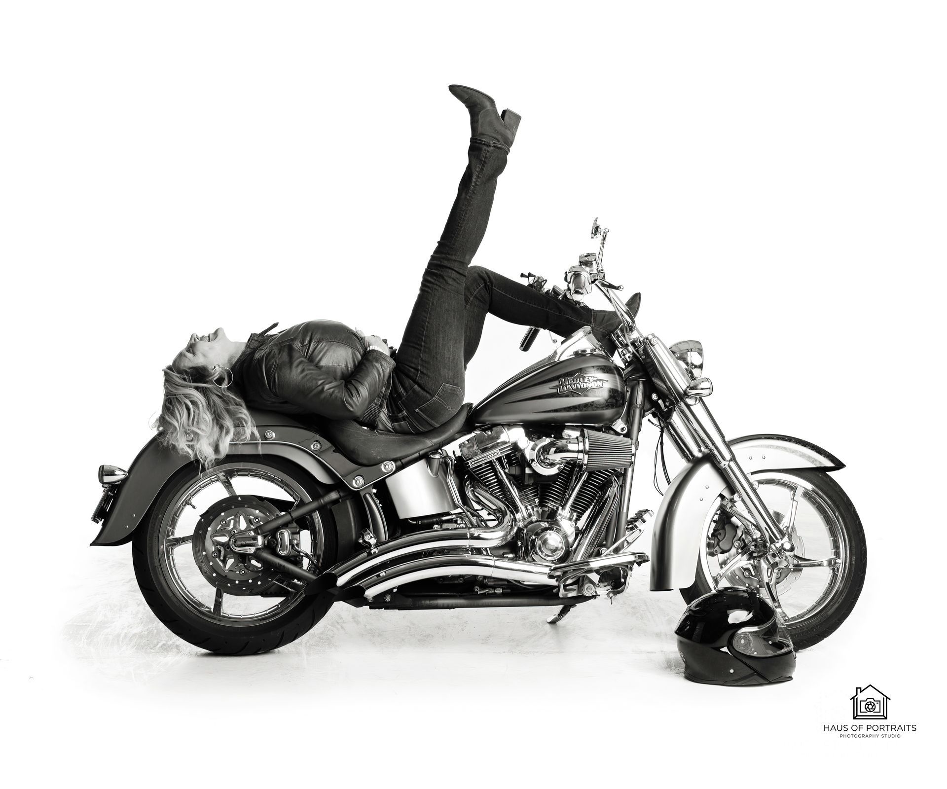 Person in black leather clothes lying across a silver motorcycle with one leg raised, studio white background