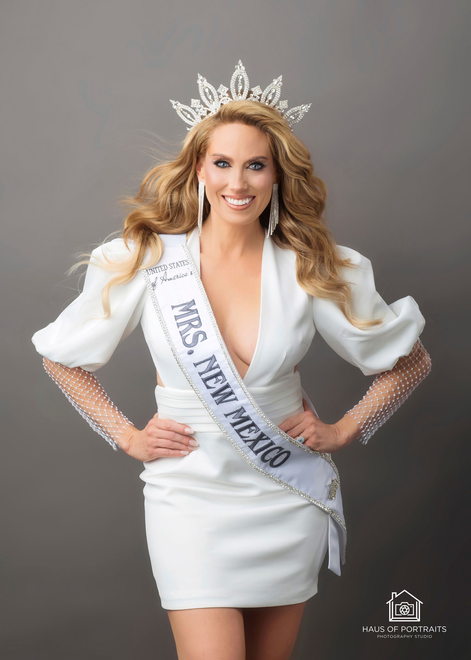 Woman in white pageant dress and crown, wearing sash, posing against a gray background.