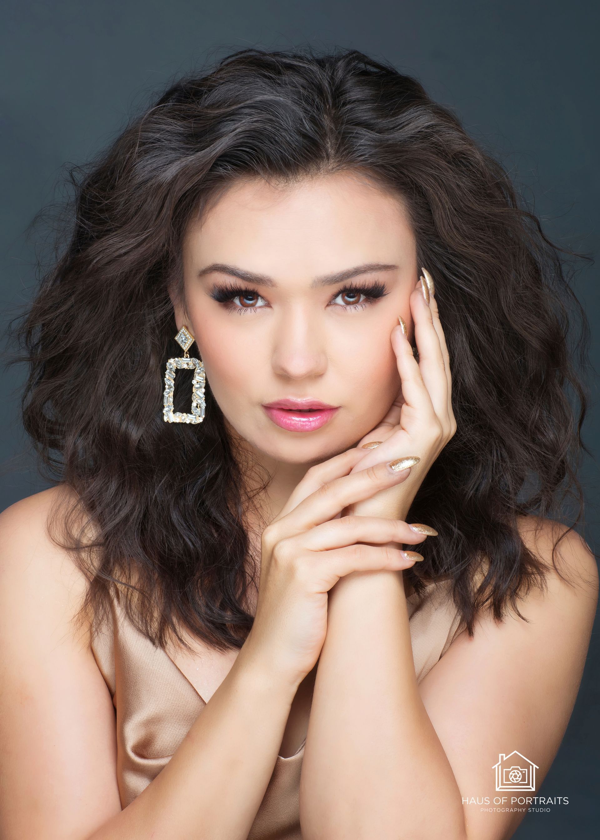 Portrait of a woman with voluminous dark hair, resting her face on her hands, wearing large earrings against a gray background