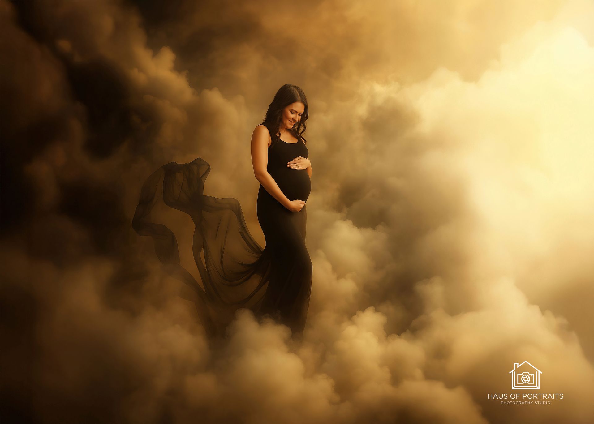 Pregnant woman in a black dress standing in golden fog with a flowing veil-like fabric
