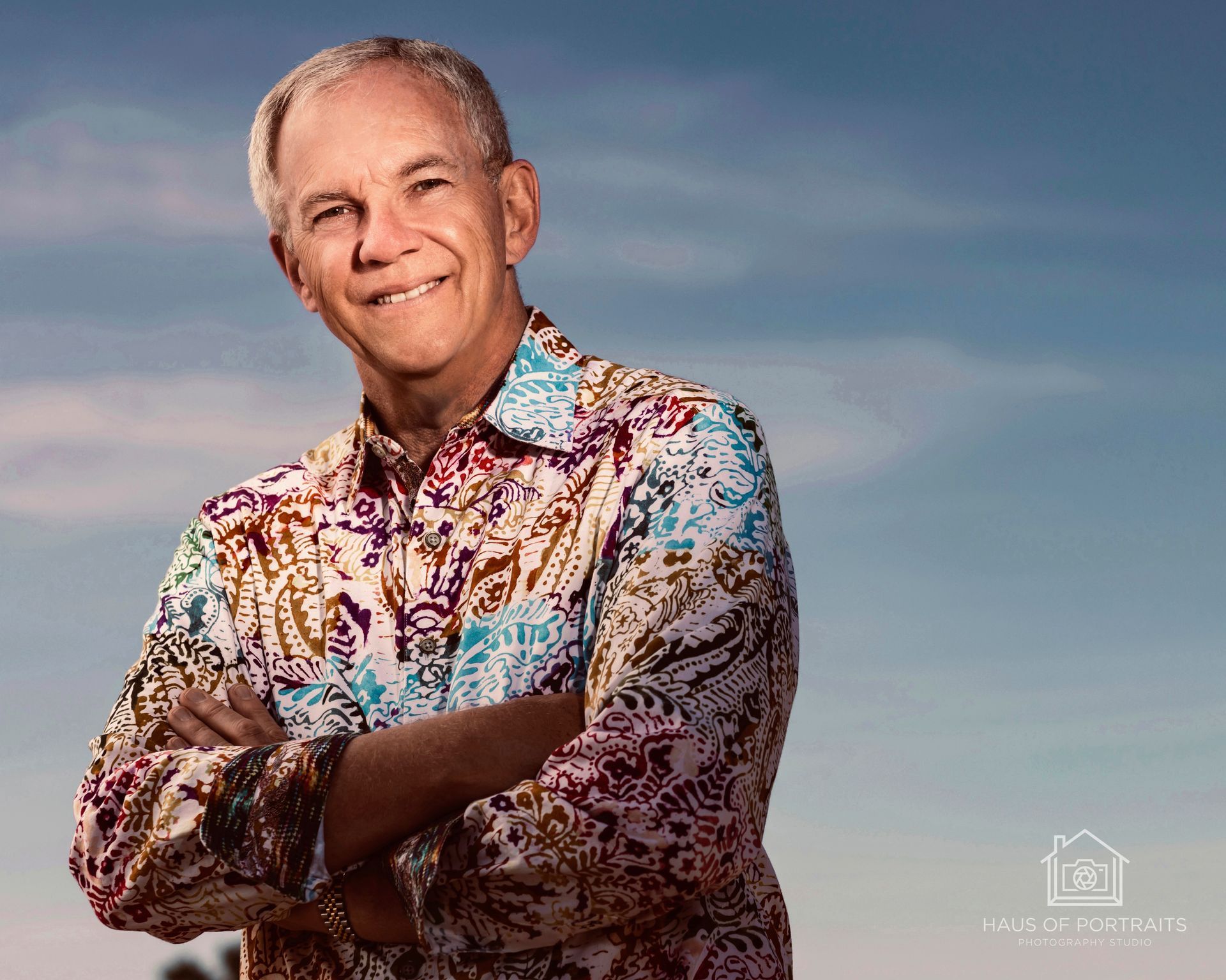 Smiling man with arms crossed in a colorful floral shirt against a sky background