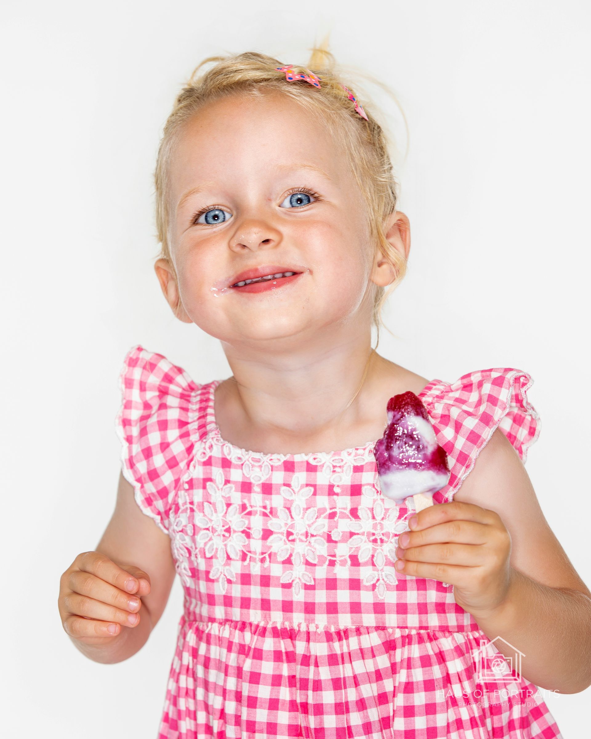 Smiling toddler in a pink checkered dress holding a pink lollipop against a white background