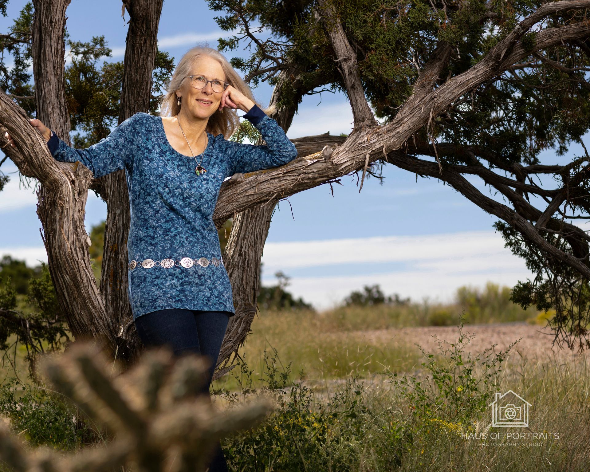 Woman in a blue sweater standing in a grassy field between large tree trunks, smiling outdoors.