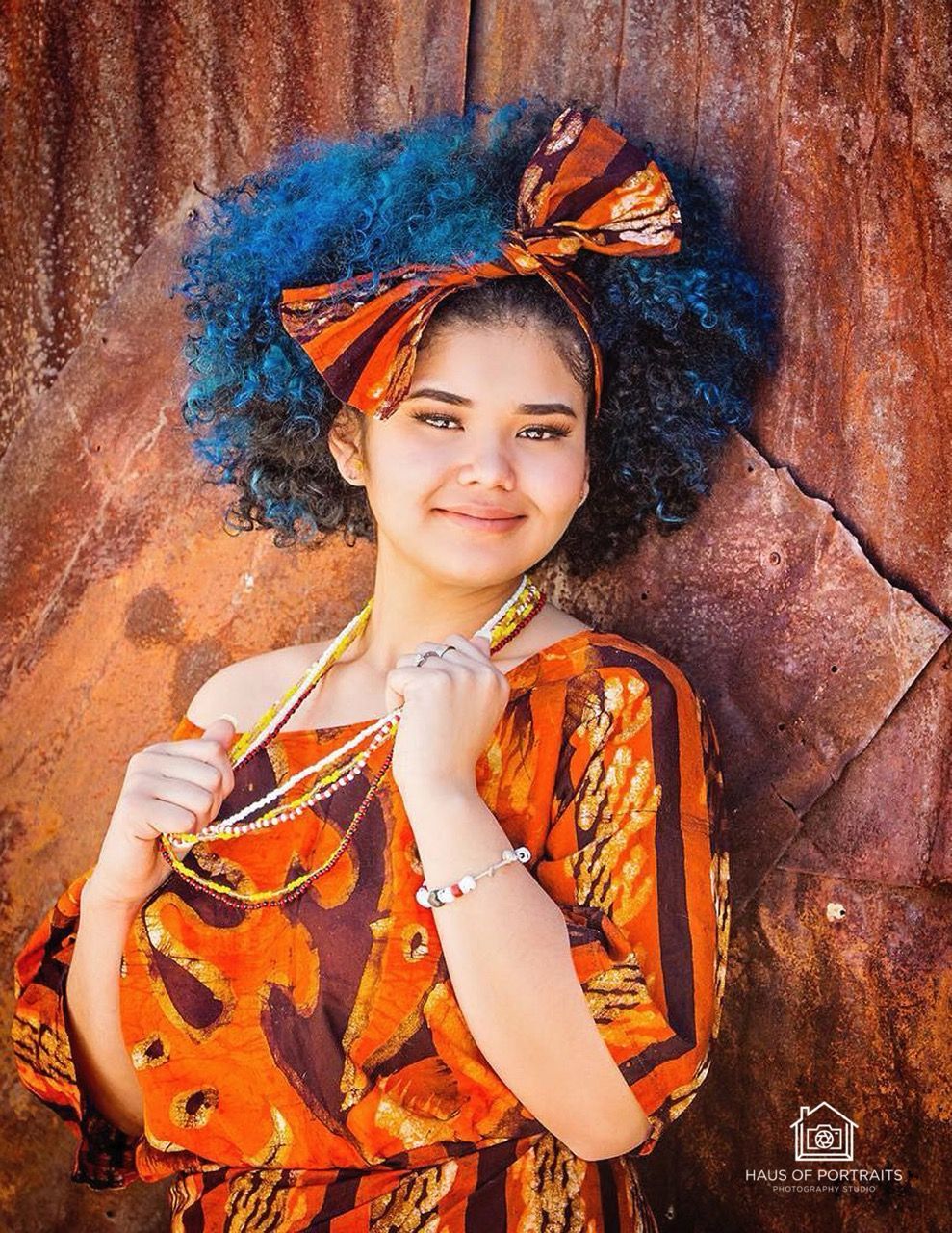 Person in orange patterned clothing and headwrap, smiling against a textured rock wall.