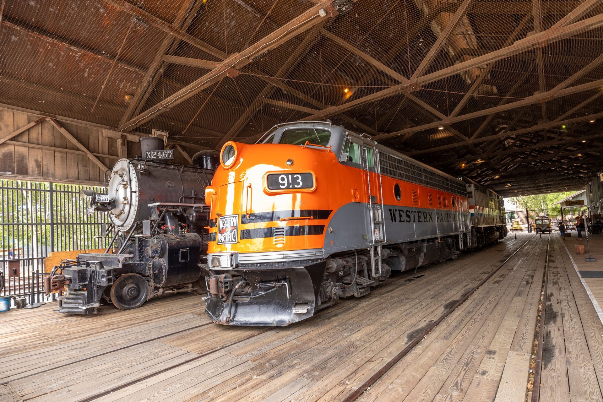 Orange and silver locomotive inside a wooden train roundhouse with another black steam engine beside it