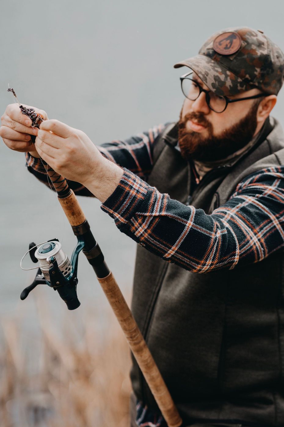 Angler holding a fishing rod and reeling in line outdoors, wearing a cap, vest, and plaid shirt.
