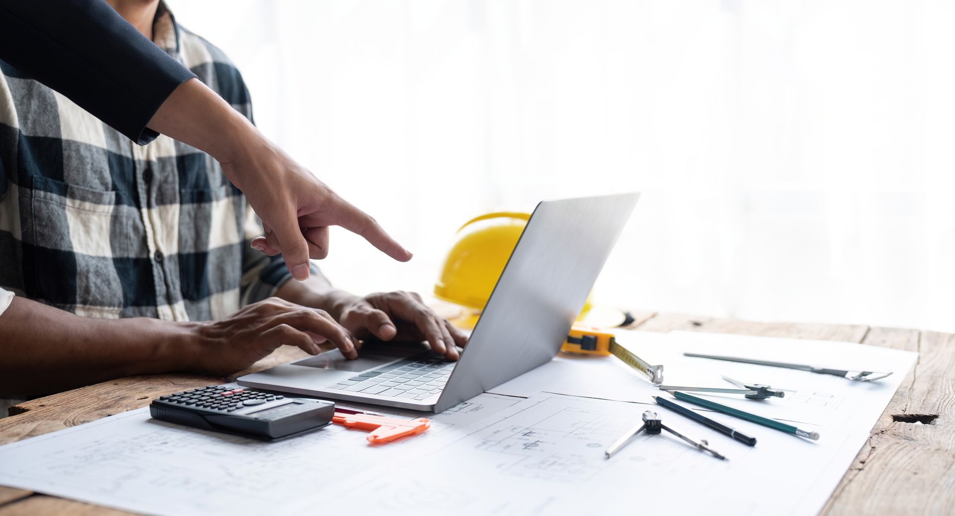 Une personne désigne l'écran d'un ordinateur portable ; des plans de construction, une calculatrice et des outils sont posés sur le bureau.