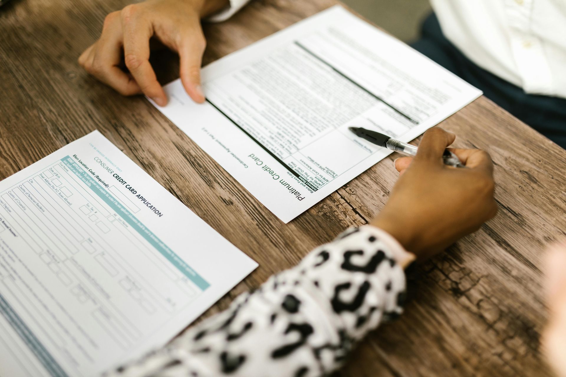 Des mains examinent des documents sur une table en bois, un stylo et des formulaires étalés dessus.