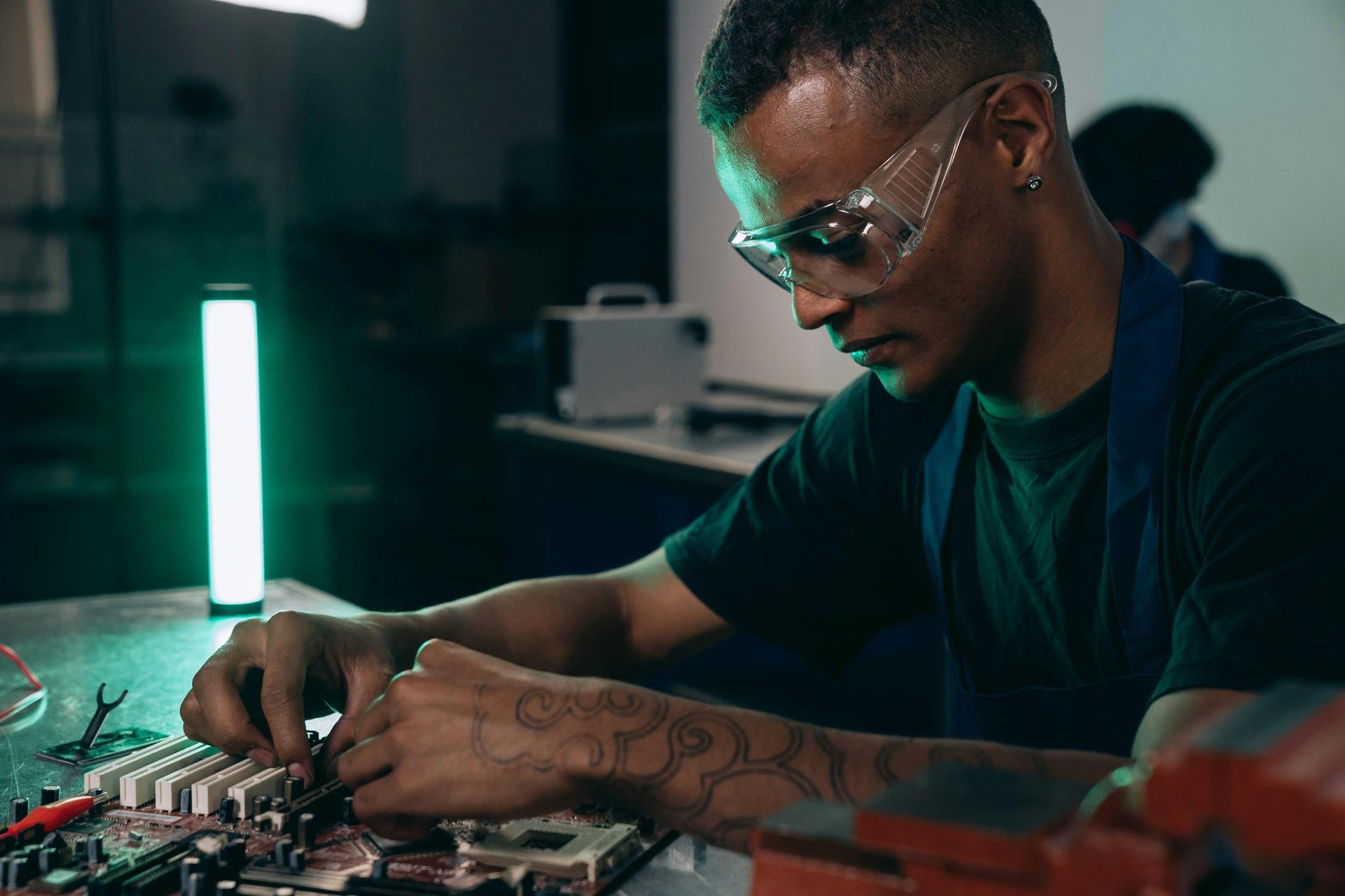 Une personne soude des composants électroniques sur un établi, éclairé par une lumière verte verticale dans un atelier faiblement éclairé.
