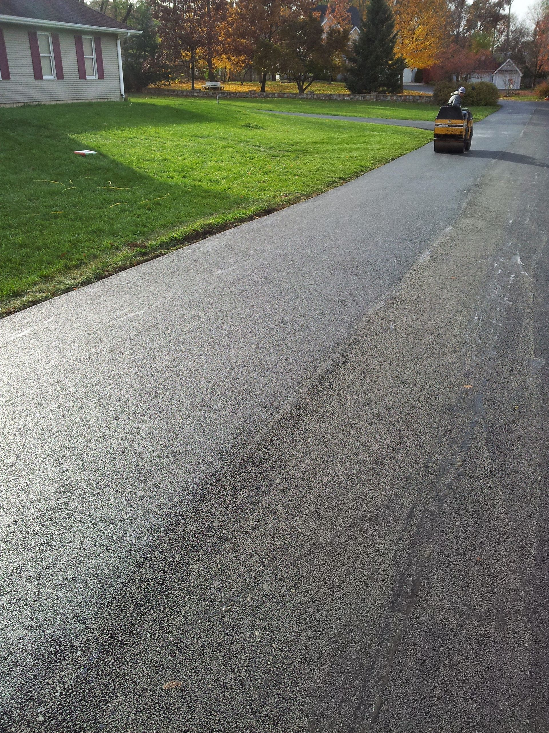 A person rides a yellow lawn mower on a paved driveway next to a grassy lawn near a house on a sunny day.