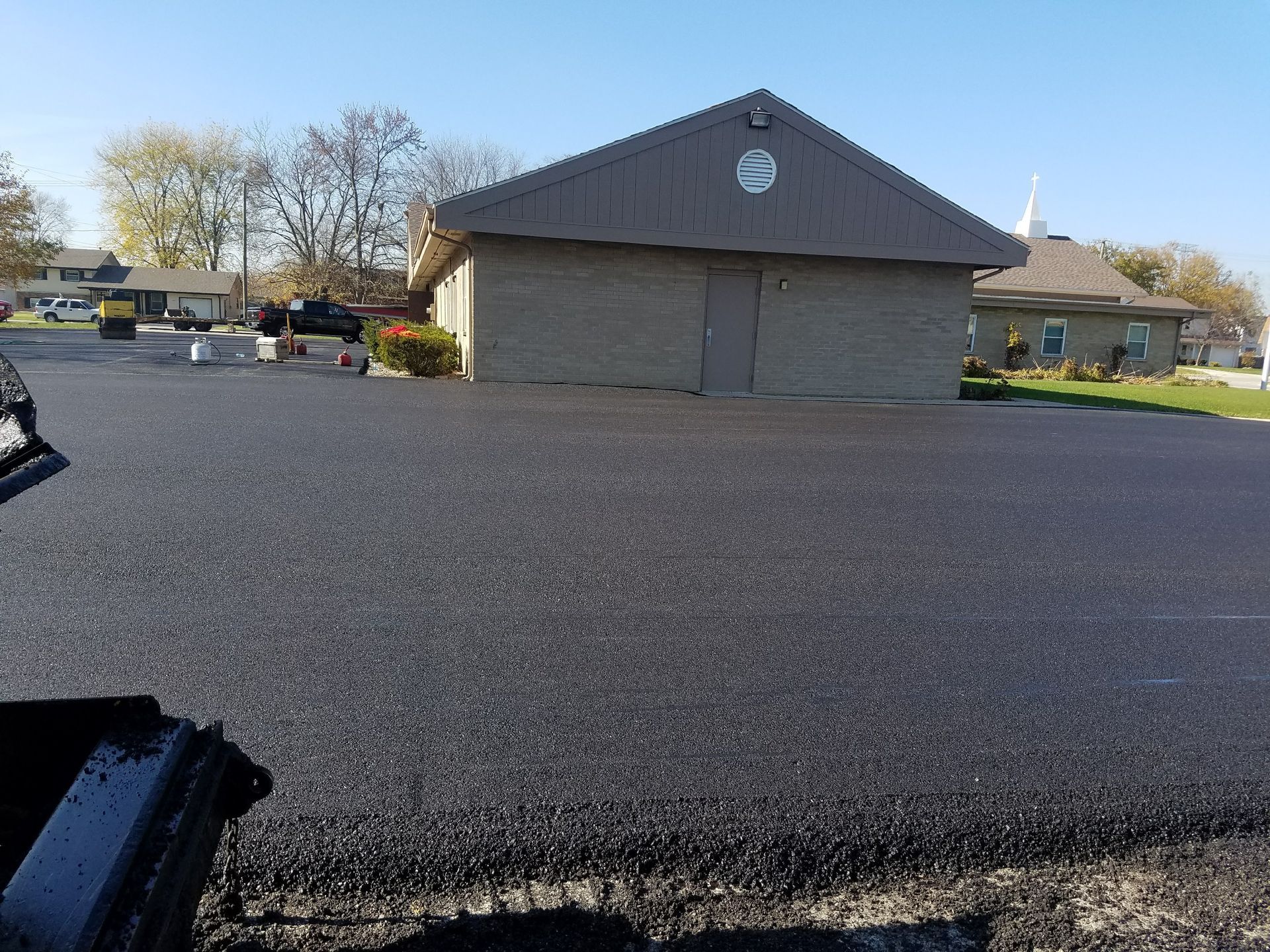 Newly paved black asphalt parking lot in front of a tan brick building on a sunny day.