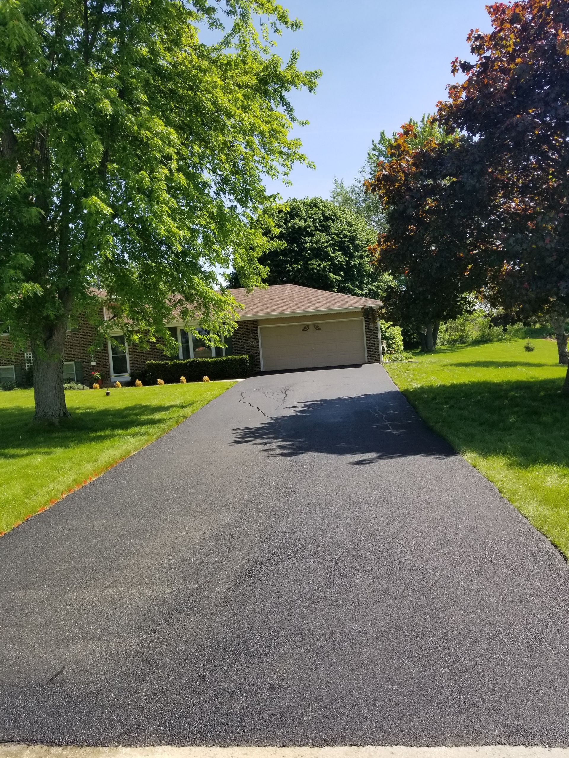 A freshly paved dark asphalt driveway leads to a suburban house with a two-car garage, surrounded by green lawns and trees.