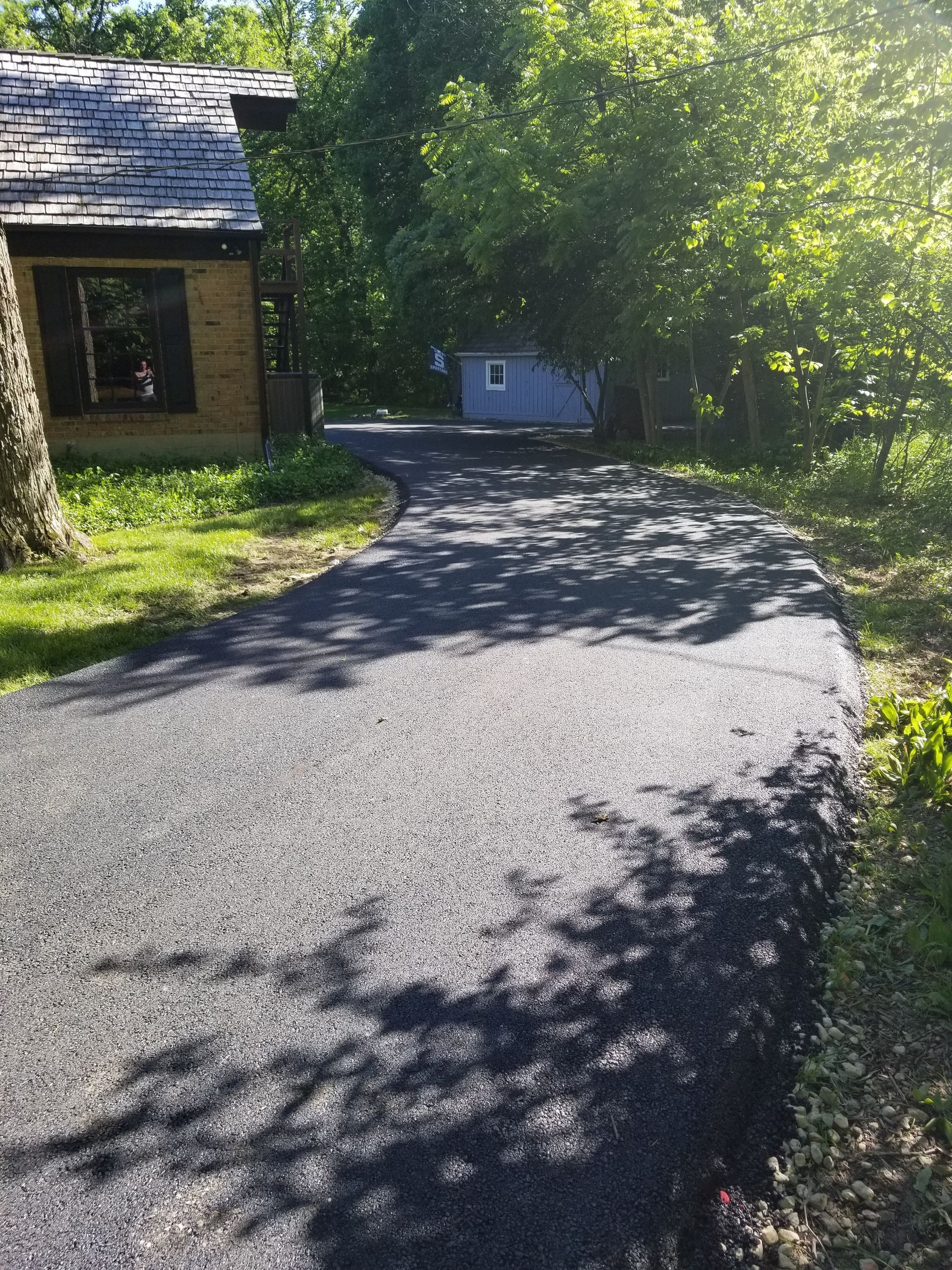 A freshly paved asphalt driveway curves through a wooded residential property toward a house and a small blue shed.