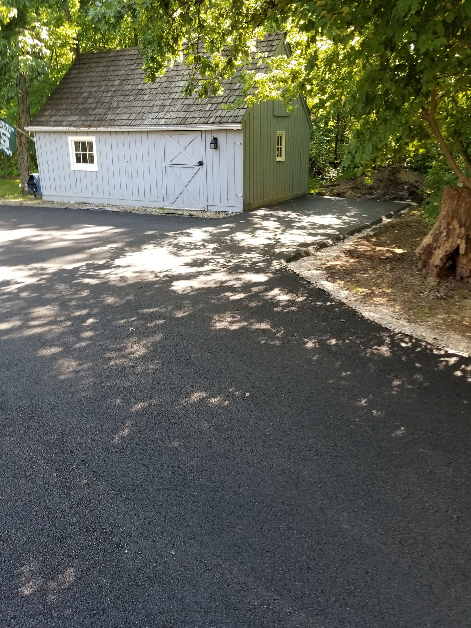 A freshly paved black asphalt driveway leads to a small, two-toned light blue and olive green shed under leafy trees.